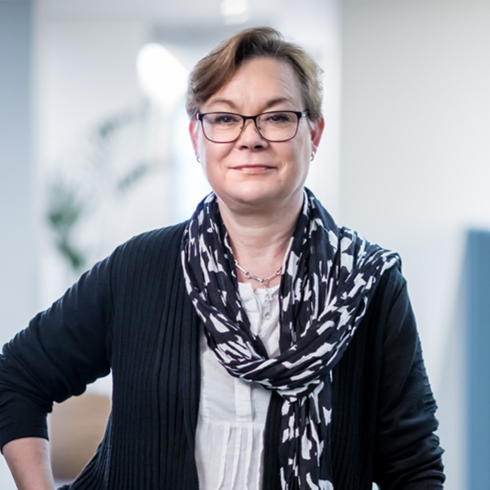 En kvinna med kort ljusbrunt hår och glasögon står inomhus, klädd i en svart kofta över en vit blus och en svartvitmönstrad scarf. Bakgrunden är mjukt suddig.