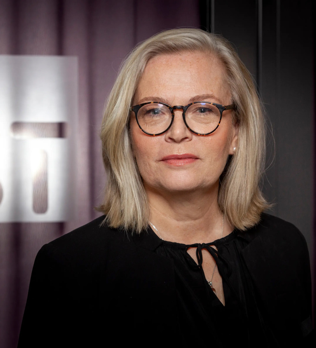 Britta Lejon, en kvinna med axellångt blont hår och glasögon ses från axlarna och upp. Hon är klädd i en svart blus med knytband i nacken. Bakgrunden innehåller en lila gardin och en delvy av en ST-skylt. Hon har ett neutralt ansiktsuttryck.