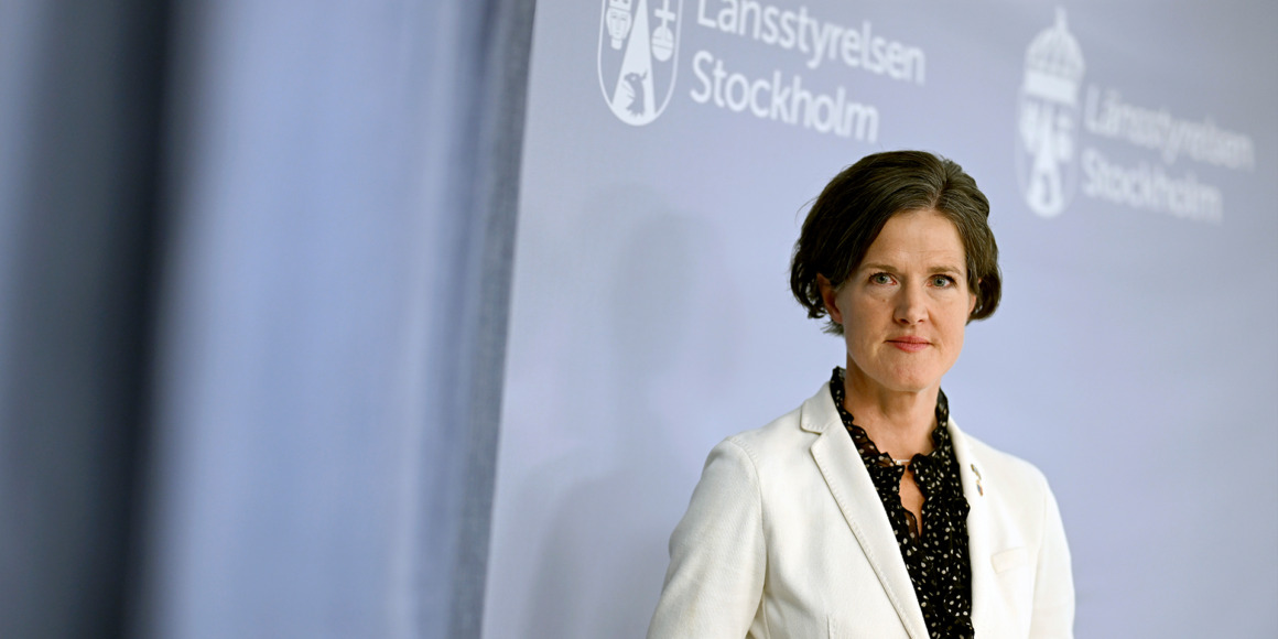 Anna Kinberg Batra, i vit kavaj och svart prickig skjorta, står självsäkert framför en grå kuliss prydd med texten "Länsstyrelsen Stockholm".