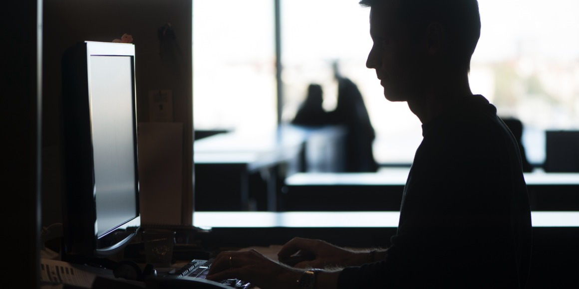 En person är silhuettad mot ett ljust fönster medan han sitter vid ett skrivbord och skriver på ett tangentbord framför en datorskärm. Kontorsmöbler och två andra figurer i bakgrunden syns svagt.