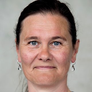 En kvinna med brunt hår tillbakadraget bär små silverfärgade örhängen. Hon har blå ögon, ljus hy och ett neutralt uttryck. Bakgrunden är vanlig och neutral-färgad.