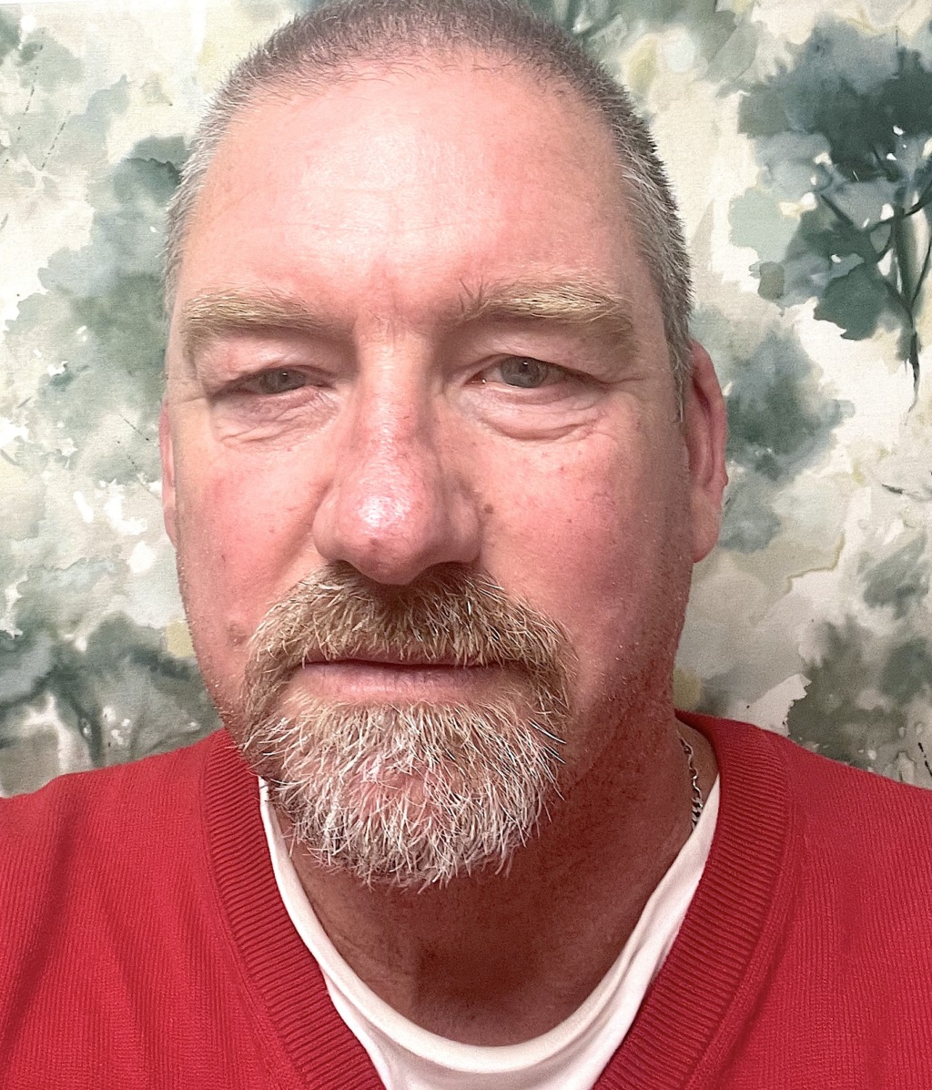 En medelålders man med kort hår och skägg ses från axlarna och upp. Han har en röd tröja på sig över en vit skjorta. Bakgrunden har ett suddigt, abstrakt akvarellliknande mönster.