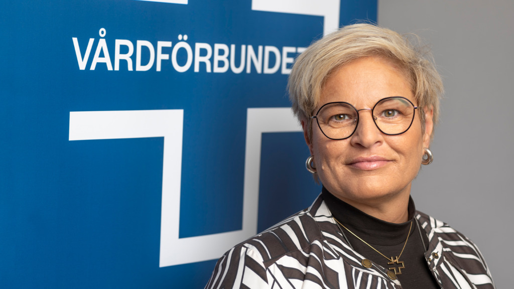 En person med kort hår och glasögon står framför en blå bakgrund med ordet "Vårdförbundet". De har på sig en randig jacka och ett halsband.