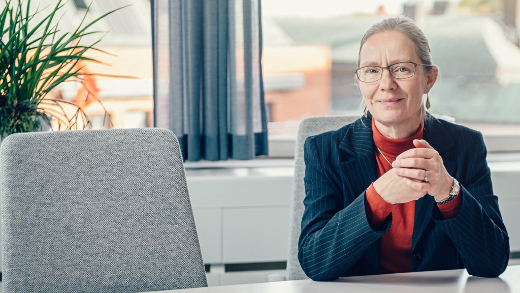 En person med glasögon och grått hår sitter vid ett bord, iklädd en svart kritrandig jacka och röd topp. Deras händer är knäppta. Det finns en växt och ett fönster med gardiner i bakgrunden.