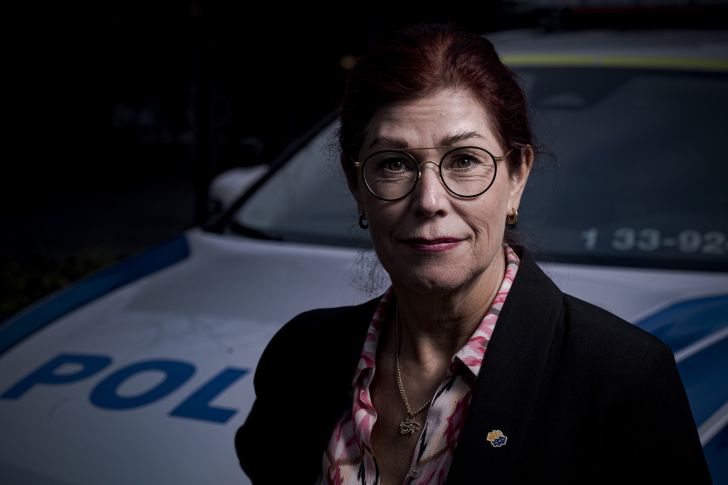 Katharina von Sydow i rött hår och glasögon som står framför en polisbil. Hon bär en mörk kavaj över en mönstrad skjorta. Bakgrunden är svagt upplyst.