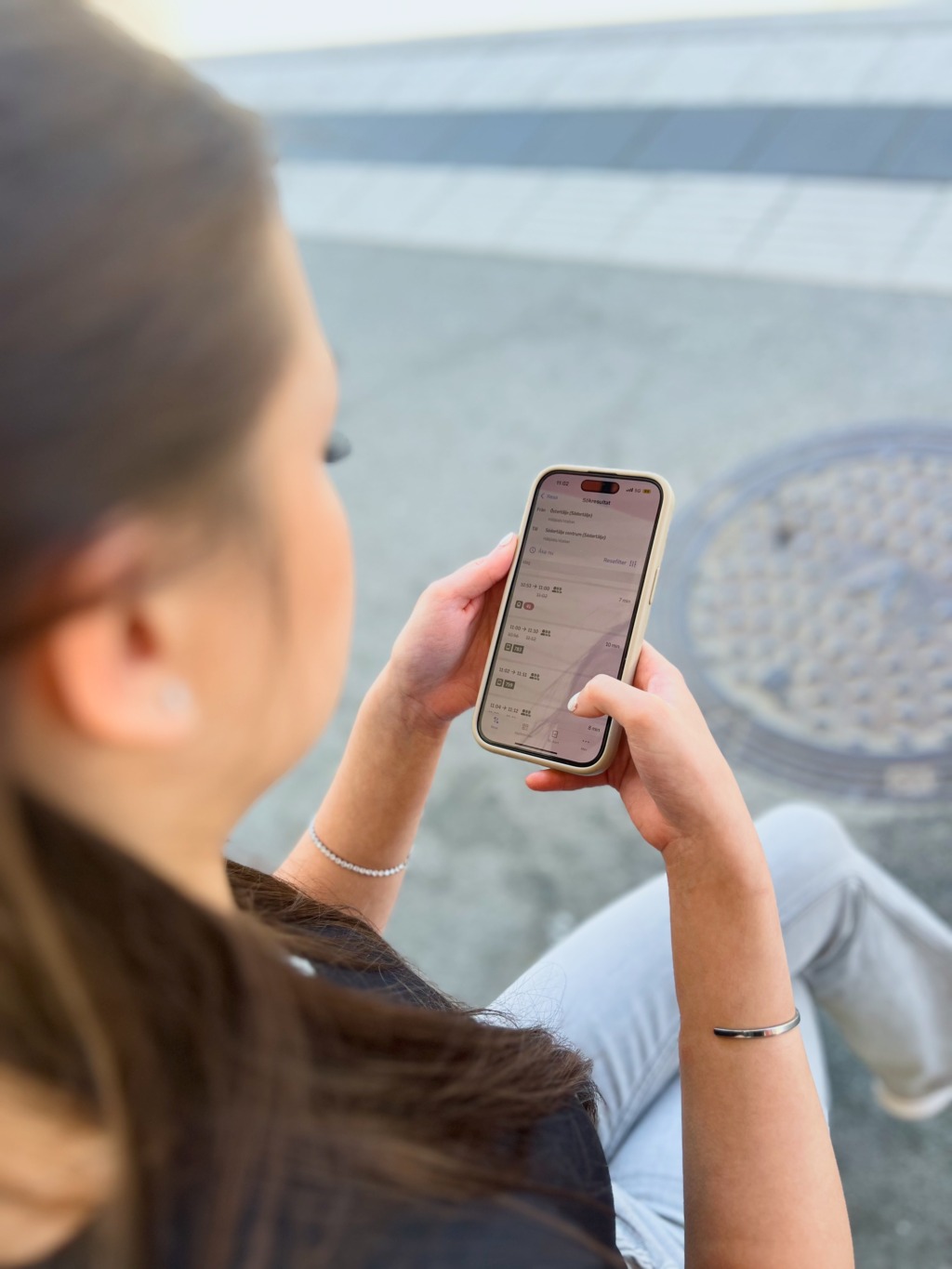 En person som sitter utomhus håller i en smartphone och använder sin högra tumme. Personen ses bakifrån och något ovanifrån, med telefonens skärm delvis synlig.