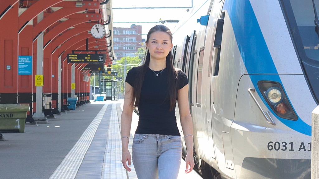 En ung kvinna med långt hår, klädd i svart skjorta och ljusa jeans, går längs en tågperrong bredvid ett stillastående blåvitt tåg i dagsljus - en scen från jobbpendling med kollektivtrafik. I bakgrunden syns skyltar och bänkar.