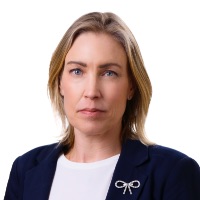 En kvinna med axellångt blont hår, klädd i en marinblå kavaj, vit topp och silverfärgad rosettformad brosch, tittar rakt in i kameran med ett neutralt uttryck - en klassisk look för professionella inom kollektivtrafik arbetspendling.