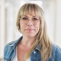 En kvinna med långt blont hår och glasögon bär en jeansjacka över en vit topp och står inomhus med ett neutralt uttryck - vilket antyder utseendet på någon som väntar under kollektivtrafik arbetspendling. Bakgrunden är mjukt suddig.