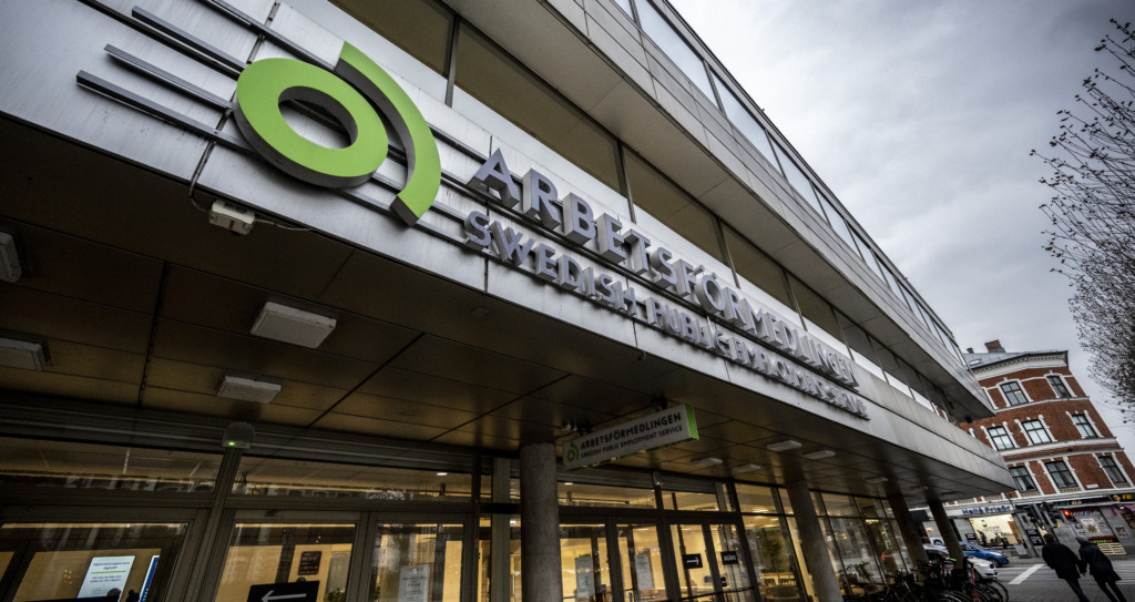 Exteriören av en kontorsbyggnad med en skylt med texten "Arbetsförmedlingen Svenska Arbetsförmedlingen" ovanför entrén, en molnig dag. Cyklar är parkerade nära entrén och några få personer syns utanför.