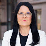 Therese Svanström, En kvinna med långt svart hår, runda glasögon och ett allvarligt uttryck bär en vit kavaj över en svart topp. Hon står framför en suddig stadsbakgrund.