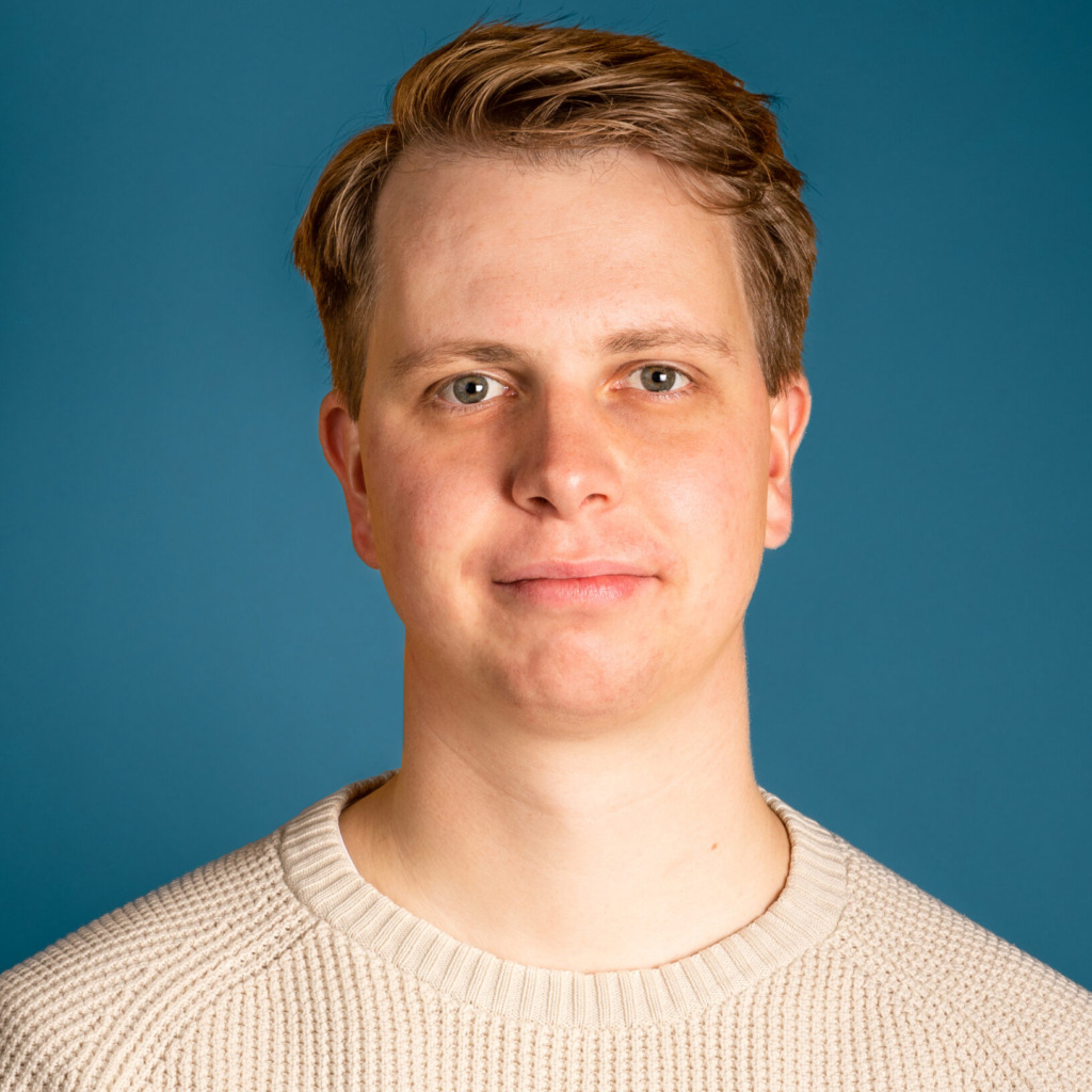 En ung vuxen med kort, ljusbrunt hår och en gräddfärgad stickad tröja står mot en solid blå bakgrund och tittar direkt in i kameran med ett neutralt uttryck, vilket förkroppsligar den minimalistiska stilen hos Oikos Timbo.