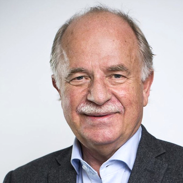 En äldre man med tunnare grått hår, mustasch och ljus hy är klädd i en blå skjorta med krage och en mörk kavaj. Han ler lätt och är vänd mot kameran mot en vanlig ljus bakgrund.