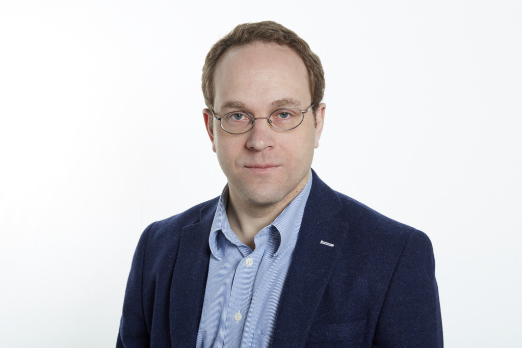 En man med kort ljusbrunt hår och glasögon, klädd i en blå skjorta och en mörkblå kavaj, står mot en vit bakgrund och tittar rakt in i kameran med ett neutralt ansiktsuttryck.