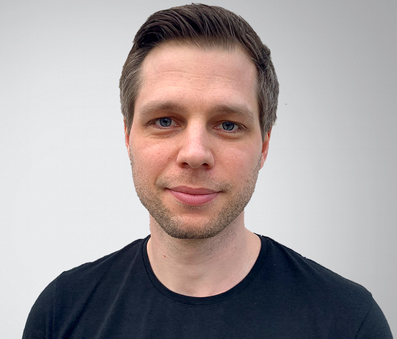 En man med kort brunt hår och lätt skäggstubb står vänd mot kameran. Han har en enfärgad svart t-shirt på sig och står mot en ljusgrå bakgrund.