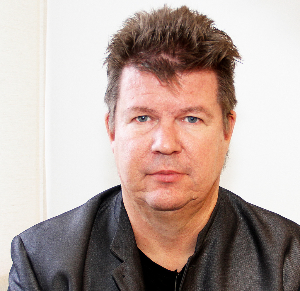 En medelålders person med kort, spetsigt brunt hår och blå ögon bär en mörk kavaj och en svart skjorta, vänd mot kameran med ett neutralt uttryck mot en slät bakgrund.