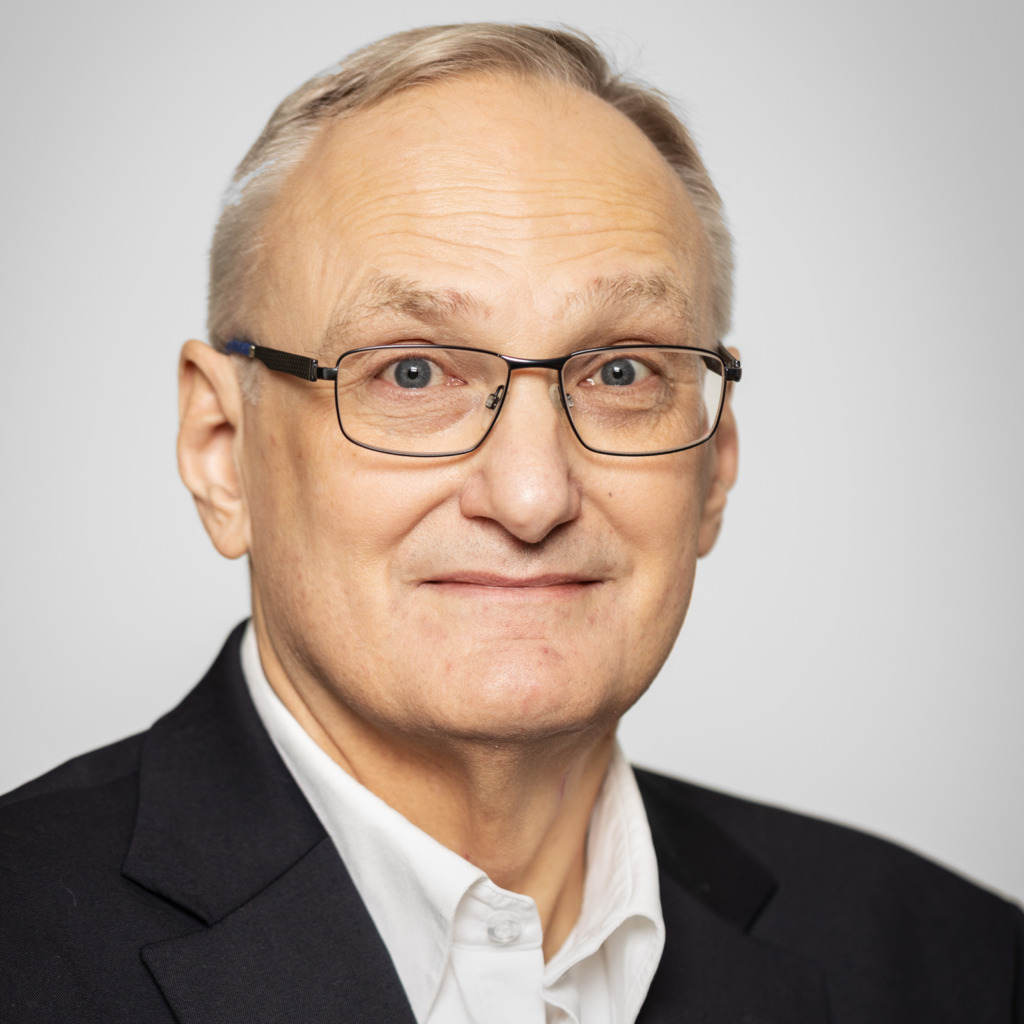 En medelålders man med kort grått hår och glasögon, klädd i mörk kostymjacka och vit skjorta, tittar in i kameran med ett neutralt uttryck mot en ljus bakgrund.