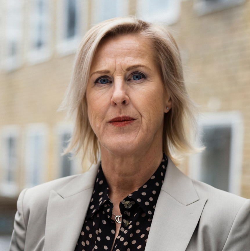 En medelålders kvinna med kort blont hår, klädd i en beige kavaj och en svart blus med vita prickar står utomhus framför en tegelbyggnad med fönster. Hon har ett neutralt ansiktsuttryck.