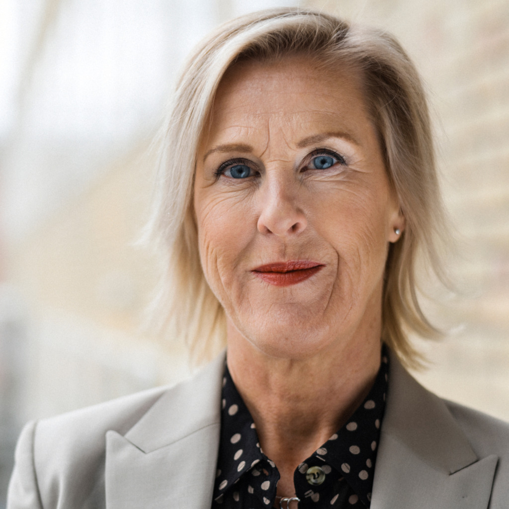 En medelålders kvinna med kort blont hår, Saco ordförande, klädd i beige kavaj och svart skjorta med vita prickar står framför en ljus tegelvägg och tittar rakt in i kameran.