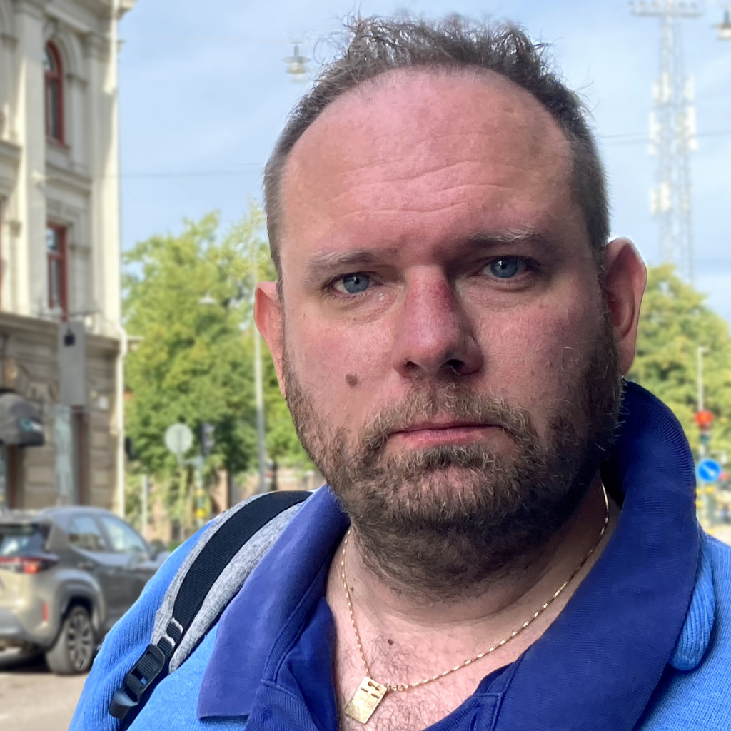 En man med kort skägg och tillbakadraget hårfäste står utomhus och reflekterar möjligen över Arbetsmiljöverkets roll i arbetet med att hindra av skyddsombud. Han bär blå skjorta, guldhalsband och ryggsäck. Byggnader, träd och parkerade bilar syns i den soliga bakgrunden.