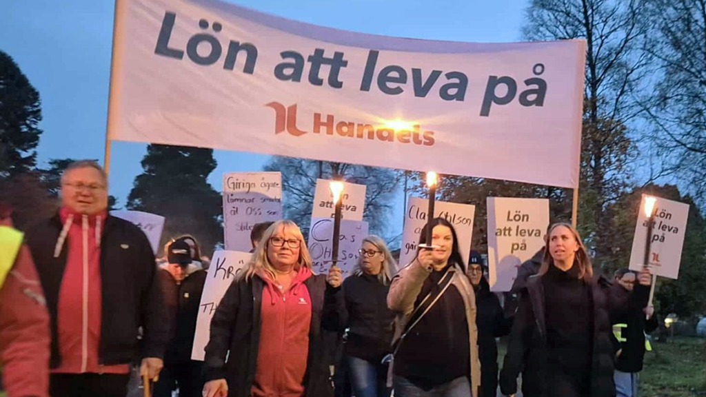 En grupp människor marscherar utomhus i skymningen med facklor och protestskyltar. På en stor banderoll längst fram står det "Lön att leva på" med Hanets logotyp. Träd och en molnig himmel syns i bakgrunden.