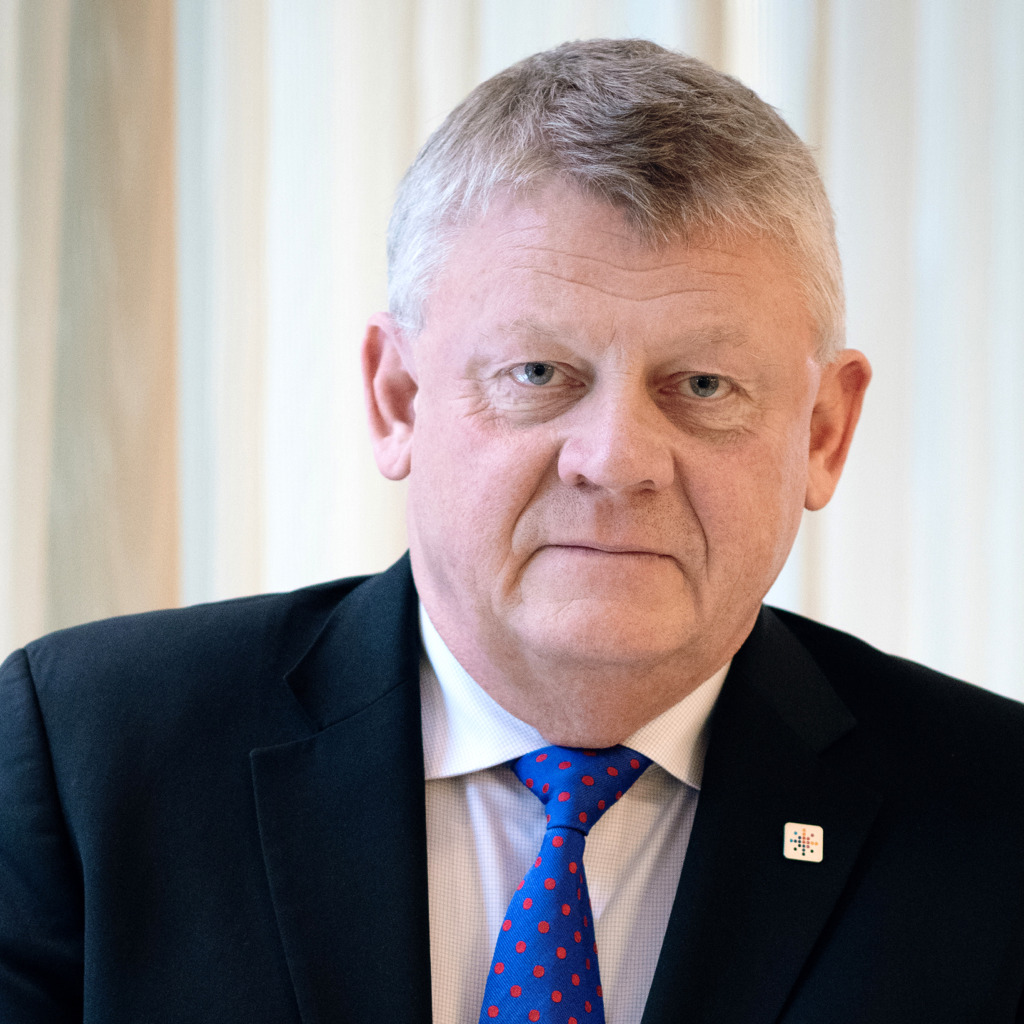 En medelålders man med kort grått hår klädd i svart kostym, vit skjorta och blå slips med röda prickar. Han är vänd mot kameran med ett neutralt uttryck. I bakgrunden syns ljusa lamellgardiner och ett Saco kongress valberedning bakgrundskontroll tema.