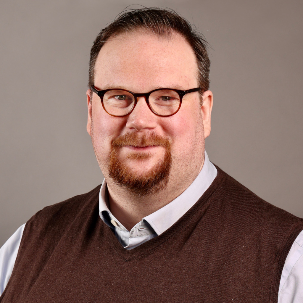 En man med kort mörkt hår, glasögon och pipskägg är klädd i ljusblå skjorta och brun tröja. Han är vänd mot kameran och ler lätt, medan nyheten om en ny offensiv mot Tesla från IF Metall gör rubriker i bakgrunden.