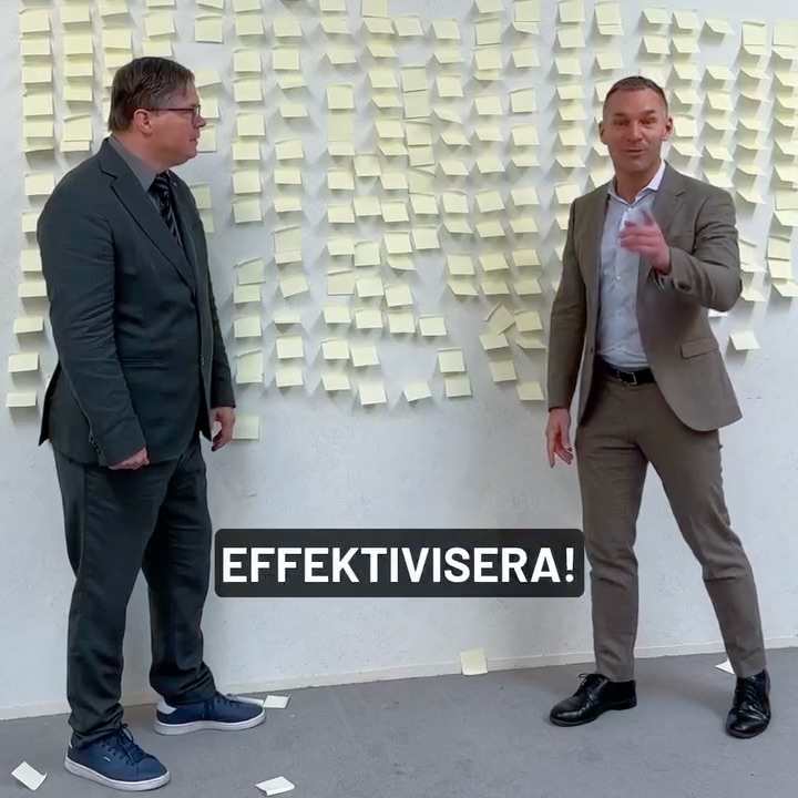 Två kostymklädda män står framför en vägg täckt av många klisterlappar. Den ena mannen gestikulerar medan den andra tittar på. Ordet "EFFEKTIVISERA!" visas i fet text längst ner i mitten av bilden.
