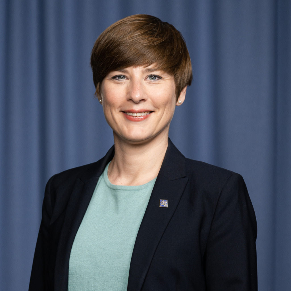 En person med kort brunt hår, klädd i svart kavaj och ljusgrön tröja, står framför en blå ridå och ler mot kameran.