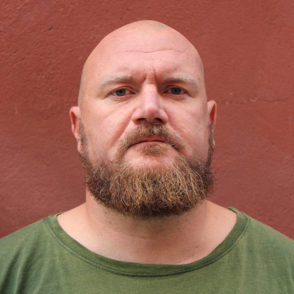 En skallig man med helskägg och mustasch i grön T-shirt står framför en slät, rödbrun vägg och tittar rakt in i kameran med ett neutralt ansiktsuttryck.