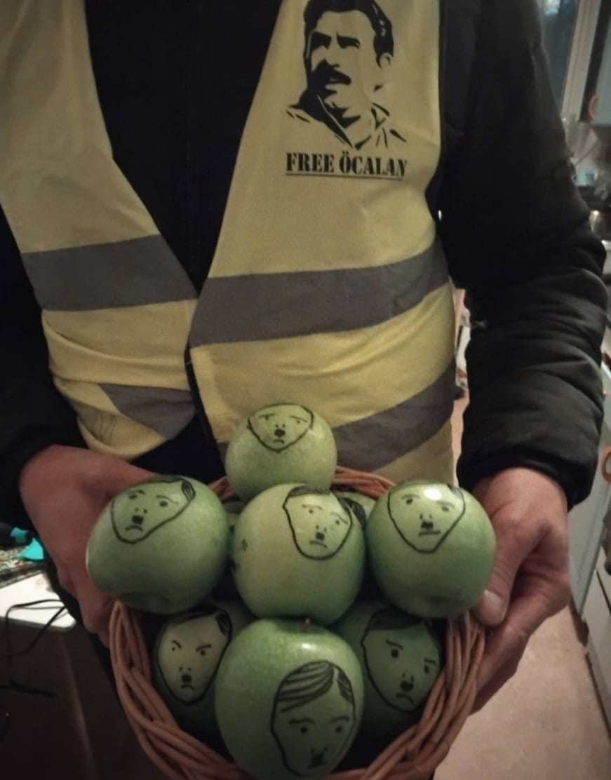 En person i gul väst med ett porträtt och texten "FREE ÖCALAN" håller en korg fylld med gröna äpplen, varje äpple har ett enkelt ansikte tecknat med svart tuschpenna som liknar Adolf Hitler.