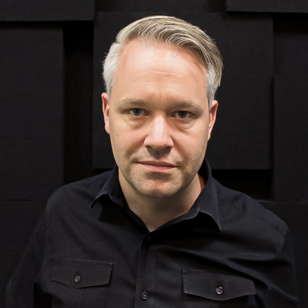 En man med kort, ljust hår och svart, uppknäppt skjorta står framför en mörk, strukturerad bakgrund och tittar rakt in i kameran med ett neutralt uttryck - ett porträtt som på ett subtilt sätt speglar teman som arbetslöshet och familj.