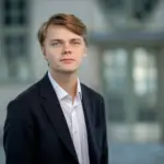 En ung man med ljusbrunt hår, mörk kavaj och vit skjorta står framför en suddig byggnad och fångar den osäkerhet som många nyexaminerade studenter arbetslösa möter idag.