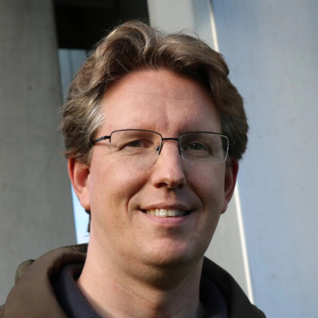 En man med ljusbrunt hår och glasögon ler mot kameran. Han bär en mörk kavaj och står framför en ljus, modern arkitektonisk bakgrund.