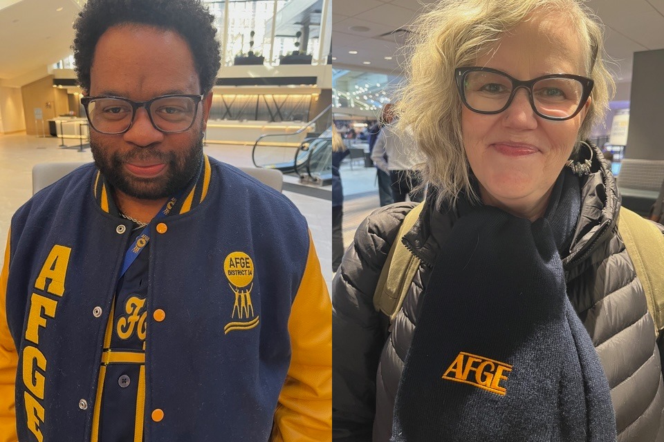 Två personer inomhus bär mörka AFGE-märkta kläder och glasögon. Den vänstra personen har en jacka med gula bokstäver och den högra personen har en halsduk med en AFGE-logotyp. En rulltrappa och flera personer syns i bakgrunden.