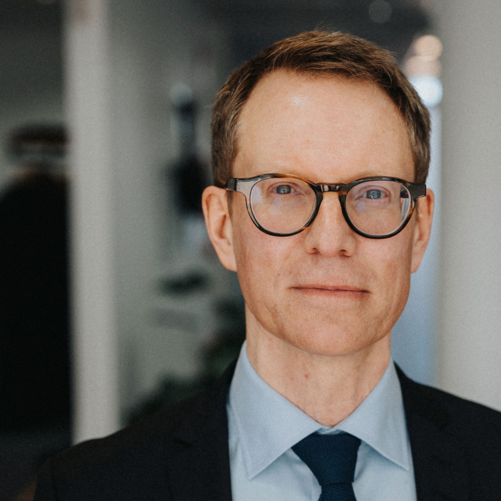 En man med kort brunt hår och glasögon, klädd i blå skjorta, blå slips och mörk kavaj, står inomhus i en svagt upplyst kontorsmiljö.