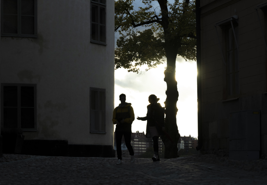 Två personer går och pratar om arbetslöshet och familj på en kullerstensgata i skymningen, deras silhuetter inramas av den glödande himlen och konturerna av byggnader och ett ensamt träd.
