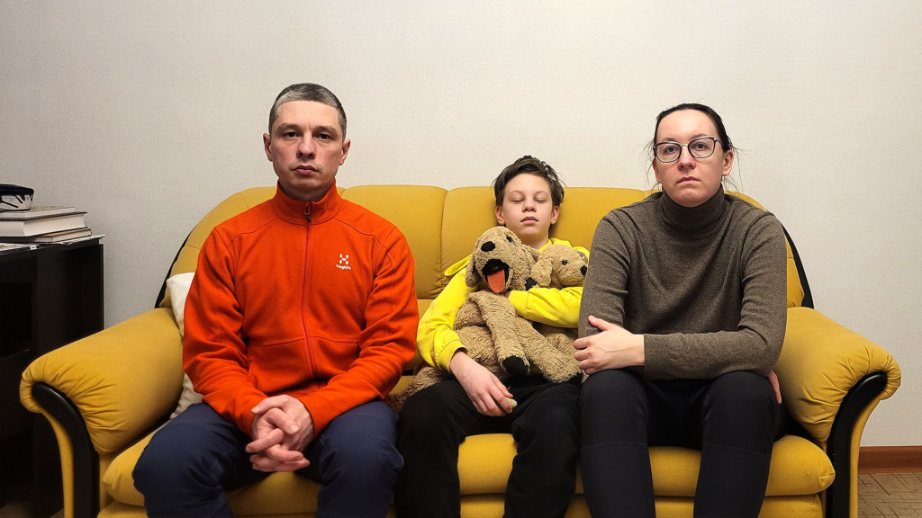 Tre personer sitter i en gul soffa mot en enfärgad vägg. En vuxen man i orange jacka sitter till vänster, ett barn med två uppstoppade hundar sitter i mitten och en vuxen kvinna med glasögon sitter till höger.