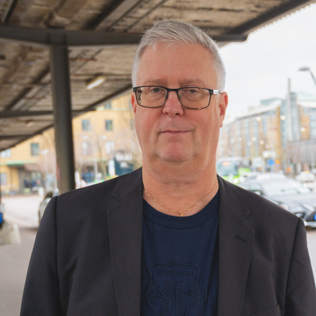 En medelålders man med kort grått hår och glasögon står utomhus under en metallkonstruktion. Han bär en mörk kavaj över en marinblå t-shirt. I bakgrunden syns byggnader, parkerade bilar och människor.