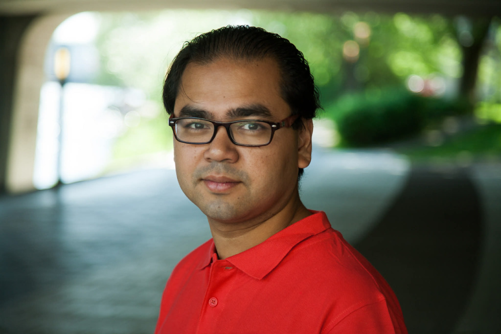 En man med kort mörkt hår och glasögon, klädd i en röd polotröja, står utomhus i ett skuggigt område med suddig grönska och solljus i bakgrunden.