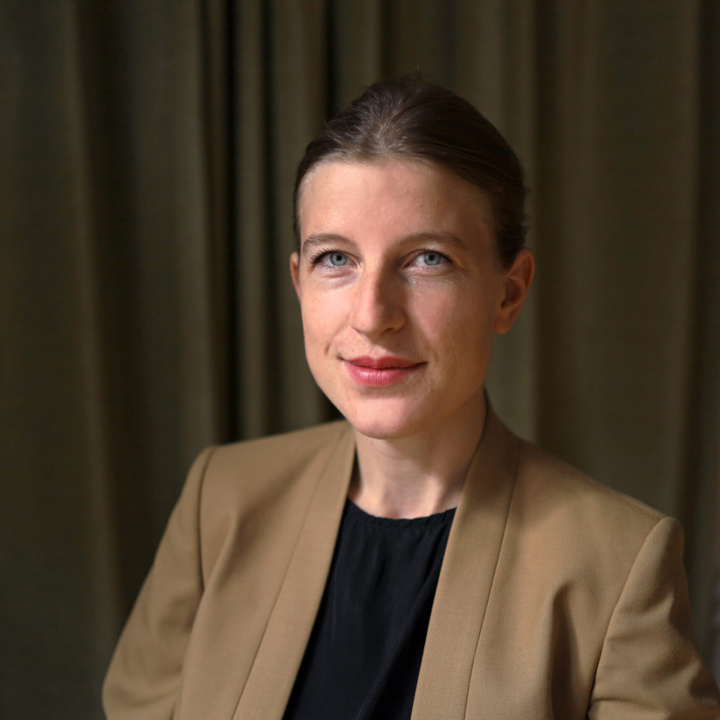 En kvinna med ljus hy och blå ögon, klädd i en brun kavaj över en svart topp, sitter framför mörka gardiner och tittar in i kameran med ett litet leende.