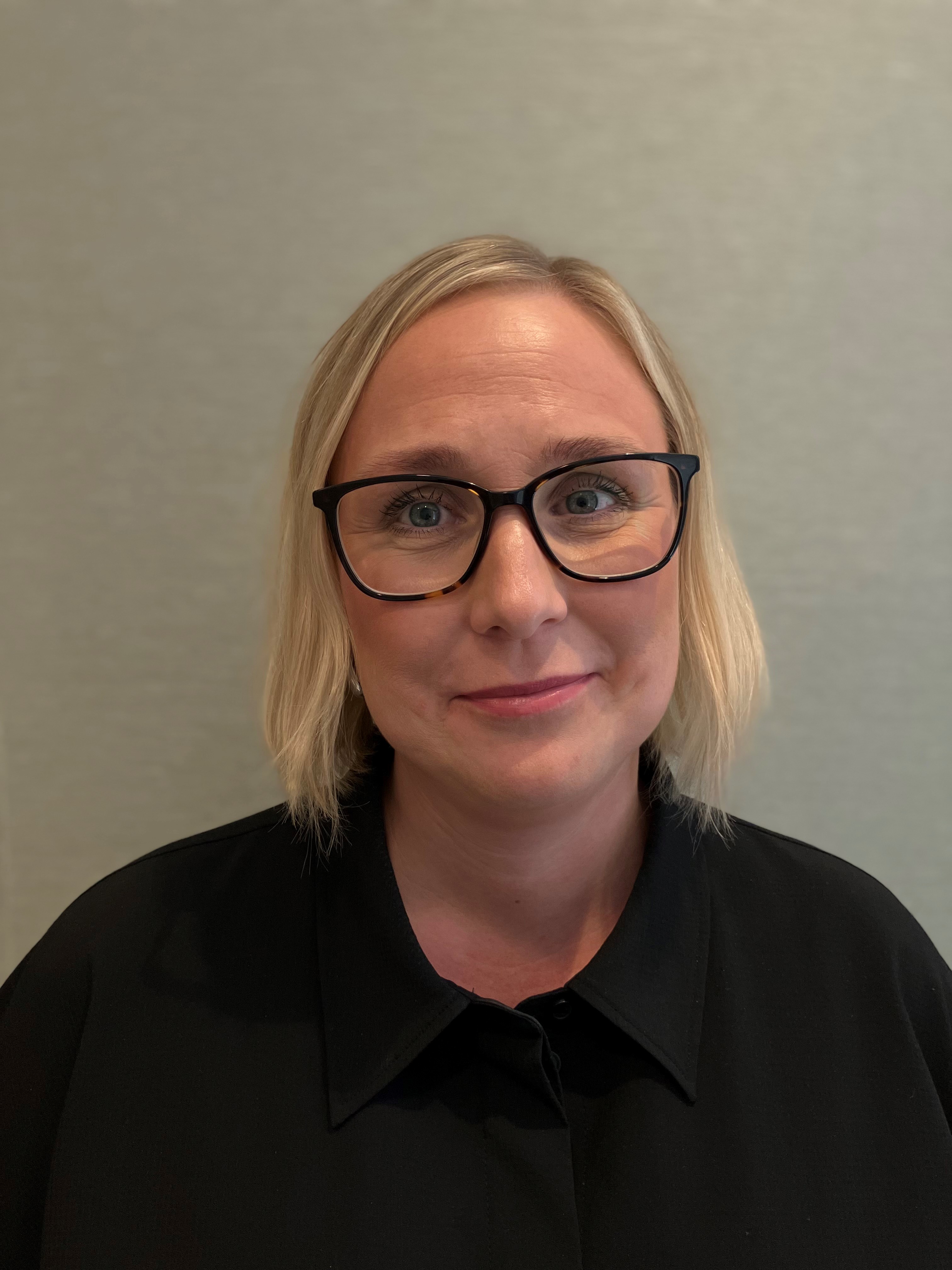 En person med kort blont hår och svarta glasögon, klädd i svart skjorta, står framför en enkel, ljus bakgrund - redo att anamma andan i Nordea kompetensskifte.