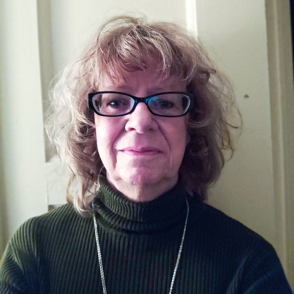 En person med lockigt, axellångt ljusbrunt hår och glasögon, klädd i en mörk ribbad polotröja och ett halsband, står inomhus framför en ljus dörr.