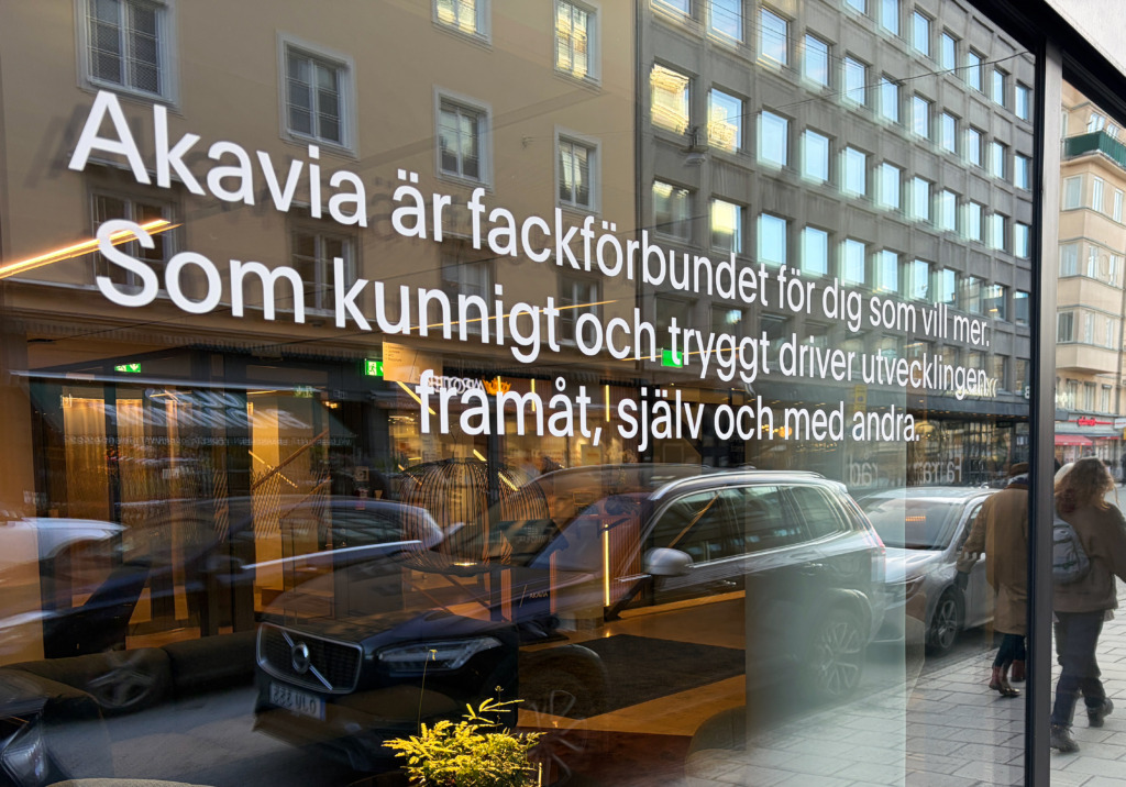En gatuscen speglad i ett glasfönster med svensk text som lyder: "Akavia är fackförbundet för dig som vill mer. Som kunnigt och tryggt driver utvecklingen framåt, själv och med andra.