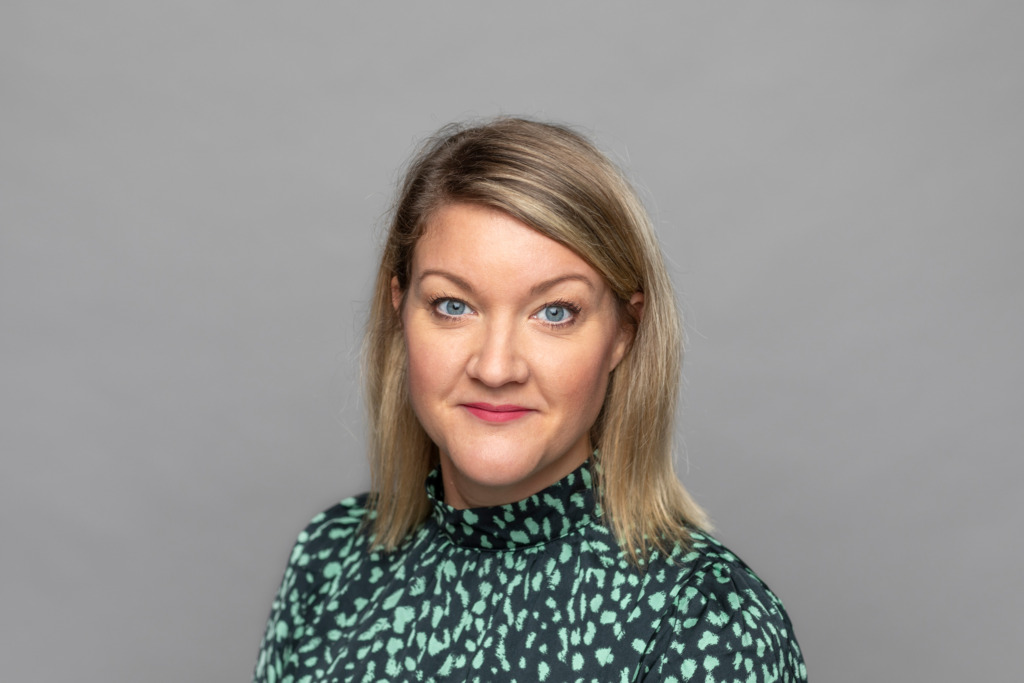 En kvinna med axellångt blont hår och blå ögon, klädd i en grön topp med svartvitt mönster, poserar framför en enfärgad grå bakgrund.