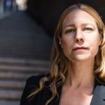 En kvinna med långt blont hår och neutralt ansiktsuttryck står utomhus nära en trappa med tegelväggar, iklädd en svart jacka. Solljuset framhäver hennes ansikte medan bakgrunden förblir dunkel.