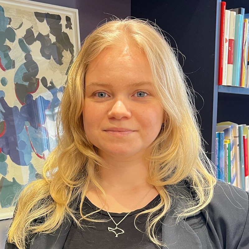 En person med långt blont hår, svart topp och jacka står inomhus, framför färgglad abstrakt konst och bokhyllor, och funderar på att välja bort barn för klimatets skull.