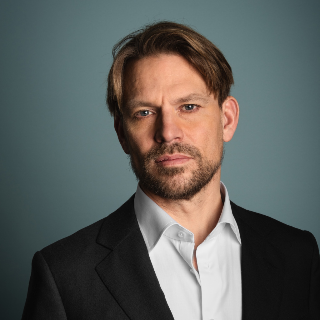 En man med ljusbrunt hår och kort skägg bär en svart kostymjacka och vit skjorta och tittar direkt in i kameran med ett neutralt uttryck mot en grå bakgrund.