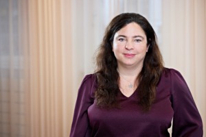 En kvinna med långt brunt hår och lila blus står inomhus framför neutralfärgade gardiner. Hon tittar in i kameran med ett lätt leende.