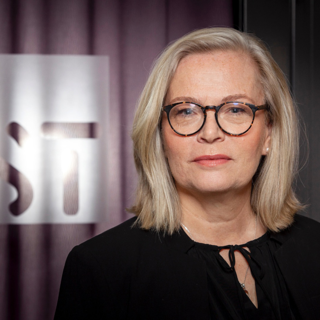 En kvinna med axellångt blont hår och runda glasögon står framför en suddig bakgrund med bokstäverna "ST" delvis synliga. Hon bär en svart topp och har ett neutralt ansiktsuttryck.